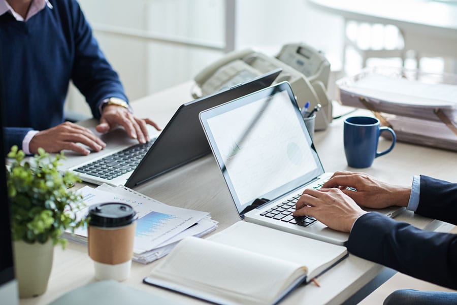 Two professionals working on laptops in a modern office setting, surrounded by documents, a coffee cup, and a plant, representing collaboration in real estate title services.
