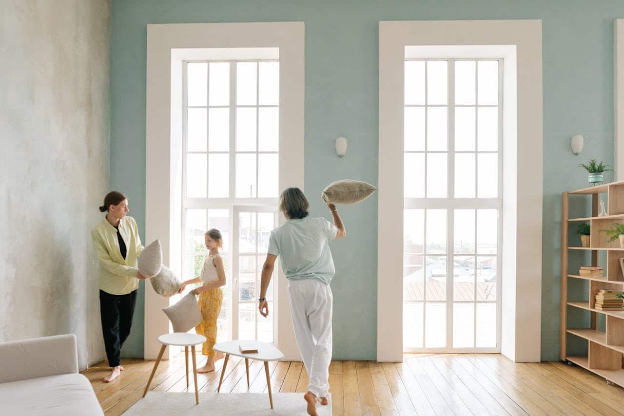 Family interacting in a bright, modern living room, with a woman handing a pillow to a girl while a man playfully prepares to toss another pillow, showcasing a warm home environment.