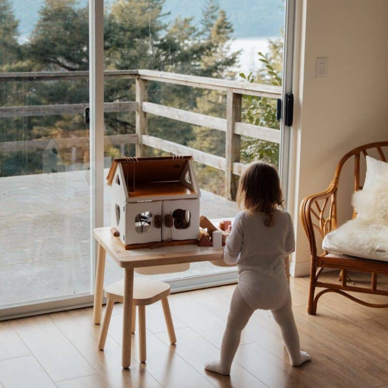 Child playing with a toy house on a table in a bright room with large windows overlooking a wooden deck and trees.