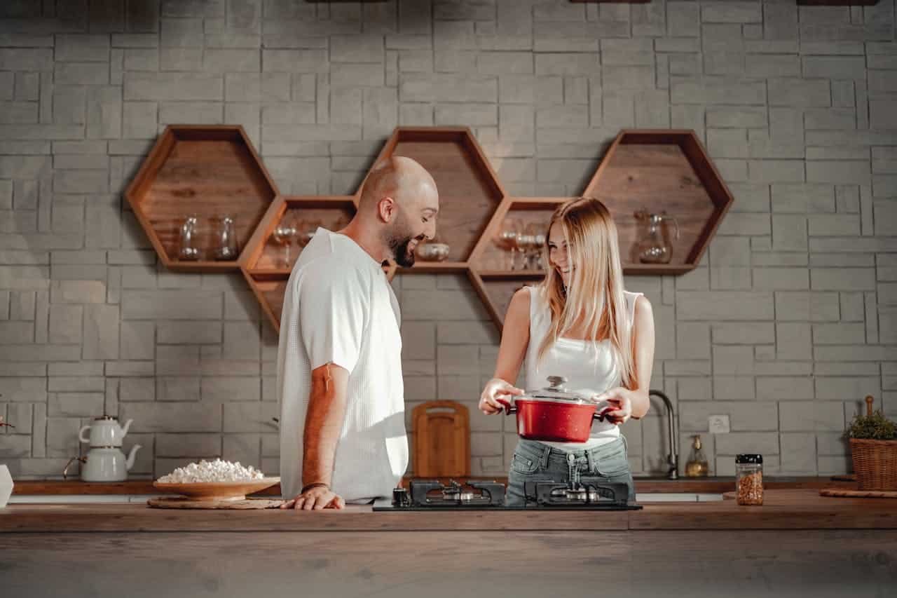 Couple in modern kitchen, man smiling at woman holding red pot, emphasizing teamwork in cooking.