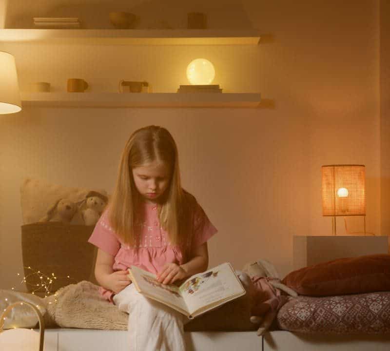 Child reading a book in a cozy, softly lit room with decorative shelves and warm lighting.