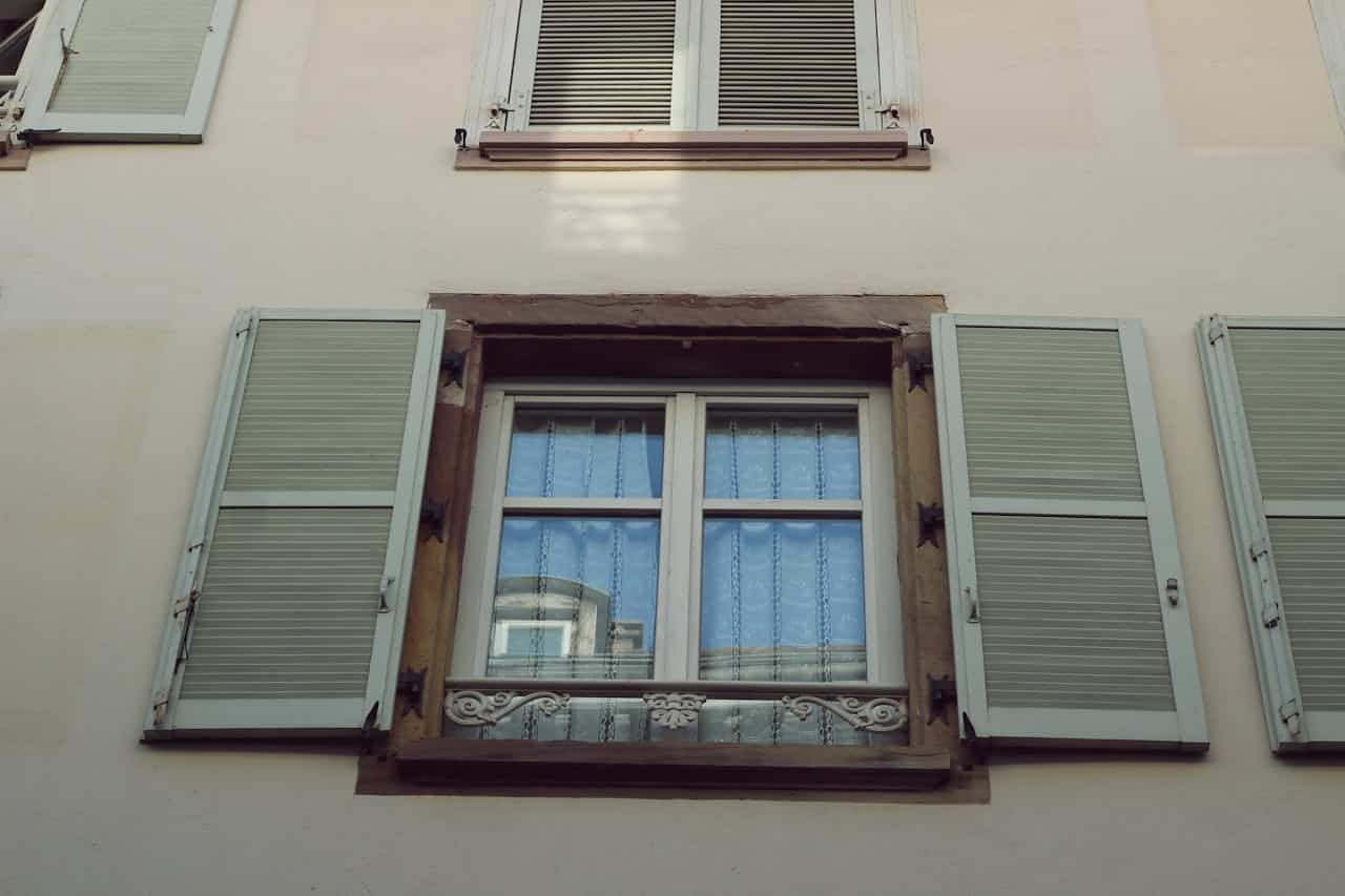 Window with decorative shutters and lace curtains on a building, illustrating architectural features relevant to real estate in Florida.