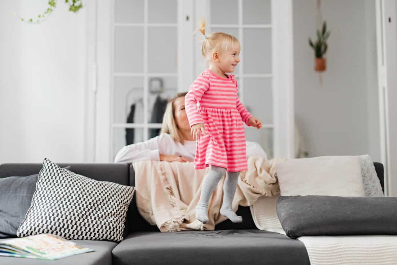 Child in pink striped dress jumping on a couch, with a parent in the background, illustrating a lively home environment.