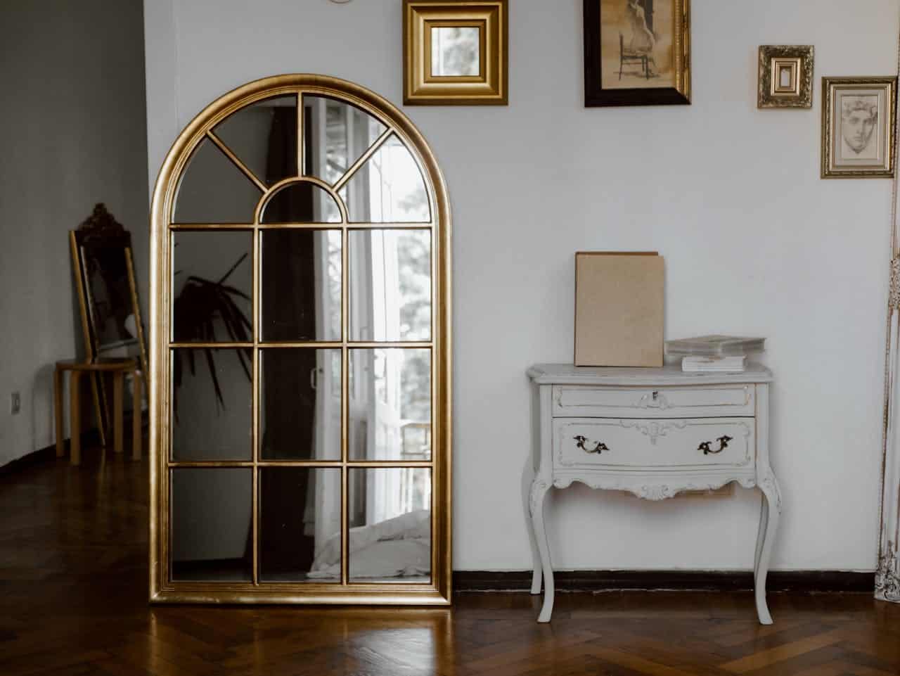 Elegant gold-framed mirror reflecting interior decor, vintage white side table with blank book and stacked papers, showcasing a stylish living space.