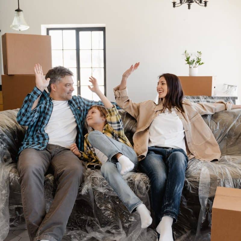 Family celebrating in a newly purchased home, sitting on a couch covered with plastic, surrounded by moving boxes, illustrating the joy of home buying and settling in.