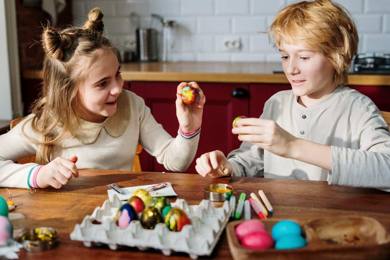 Children decorating colorful Easter eggs at a wooden table, with various art supplies and decorated eggs in view.