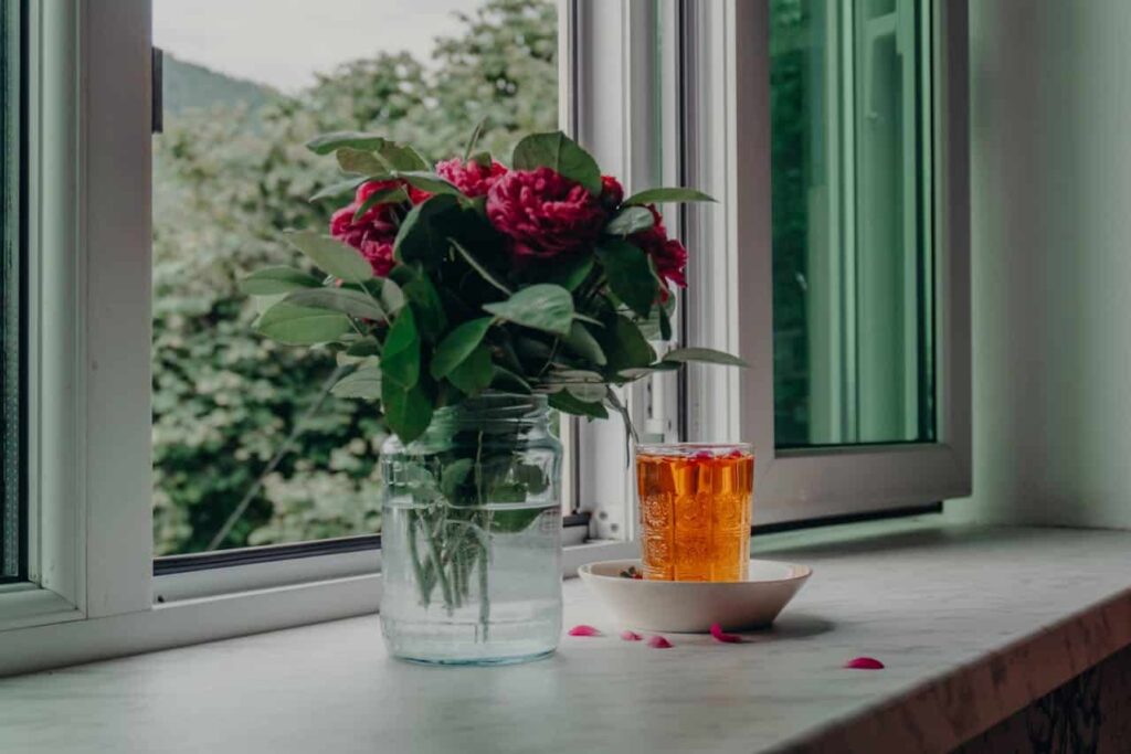 Fresh flowers in a jar and a glass of iced tea on a windowsill, surrounded by greenery, symbolizing a relaxed home atmosphere.