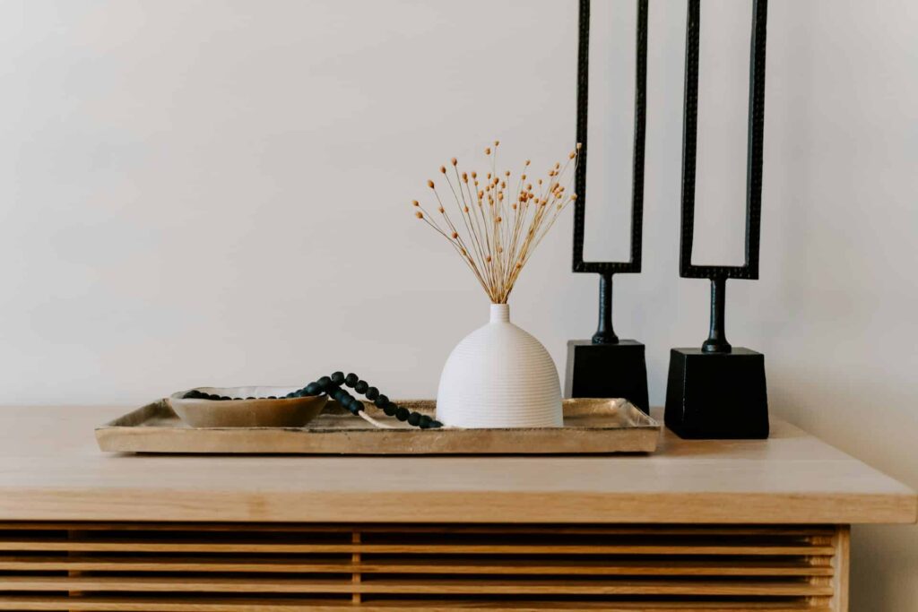 Decorative tabletop arrangement featuring a white vase with dried flowers, a small dish, and a beaded necklace, alongside black candle holders on a wooden surface.