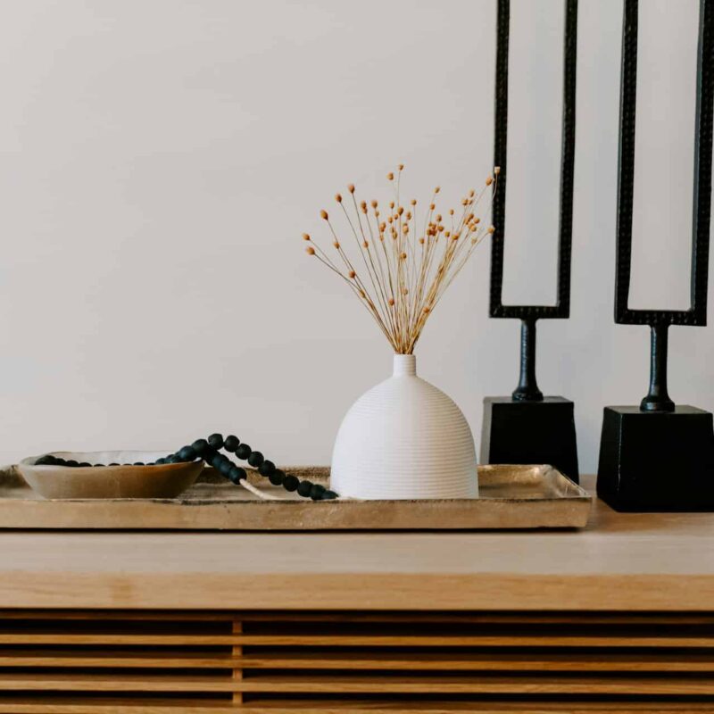 Decorative arrangement featuring a white vase with dried flowers, a black beaded necklace, and a gold bowl on a tray, alongside two black candle holders, symbolizing home aesthetics relevant to real estate and title services.