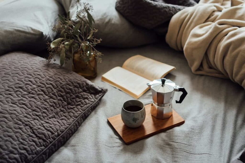 Cozy bedroom setting with pillows, a coffee pot, and a cup on a wooden tray, alongside a book and dried flowers, evoking a relaxing atmosphere for home buyers.