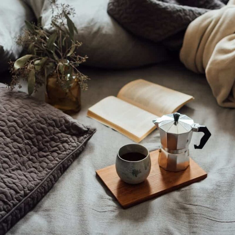Cozy scene with a moka pot, a cup of coffee, an open book, and a vase of dried flowers on a textured blanket, evoking a warm, inviting atmosphere for home buying insights.