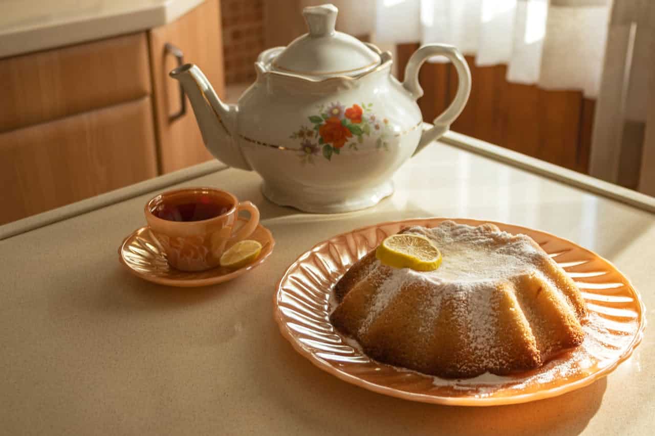 Tea set with a teapot and a cup of tea accompanied by a lemon slice, alongside a bundt cake dusted with powdered sugar on a decorative plate, set in a cozy kitchen environment.