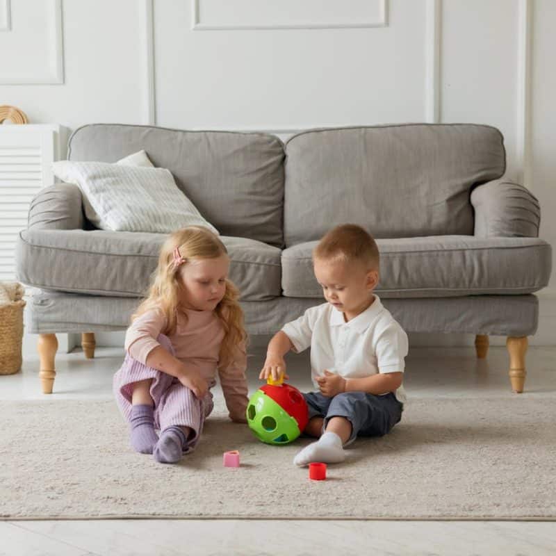 Children playing with a colorful toy ball on a soft rug in a cozy living room with a gray sofa and decorative pillows.