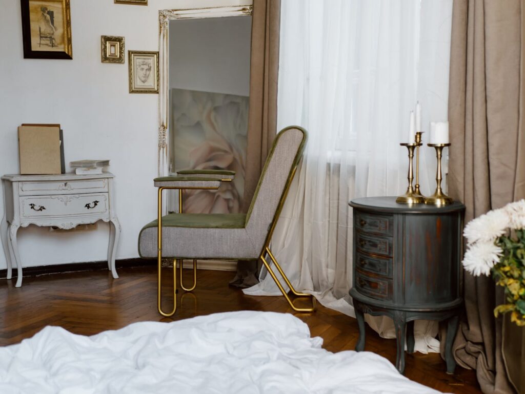 Cozy interior of a bedroom featuring a green chair with gold accents, a white bedside table with decorative items, a dark round side table with candles, and soft white bedding, reflecting a serene home environment.