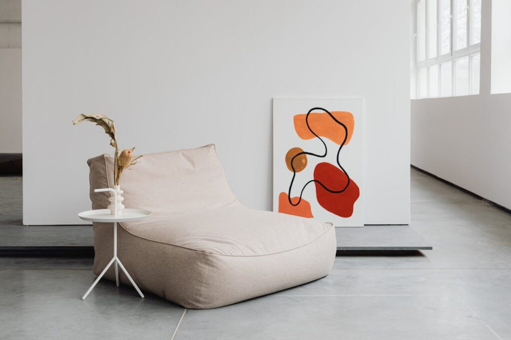 Cozy bean bag chair with minimalist design, accompanied by a small white side table holding a decorative vase, and a modern abstract painting featuring red and orange shapes on a white wall.