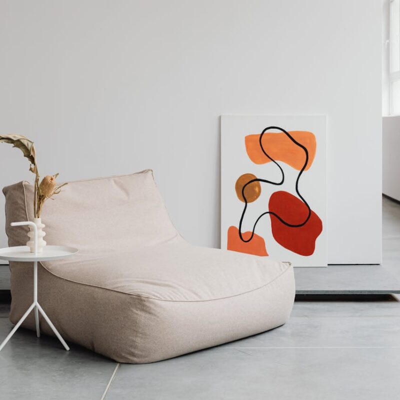 Cozy beige bean bag chair beside a small white table with a decorative vase, featuring abstract artwork in warm colors on the wall, enhancing a modern home interior aesthetic.