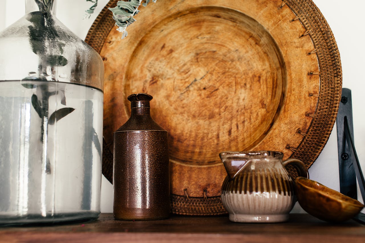 Decorative arrangement featuring a glass vase with foliage, a brown ceramic bottle, a striped ceramic jug, and a wooden platter on a shelf, representing home decor and aesthetic elements.
