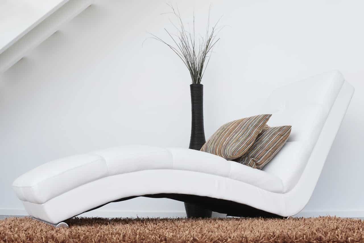 White modern chaise lounge with striped pillows and a tall black vase, set against a minimalistic white wall and brown rug, evoking a relaxed atmosphere.