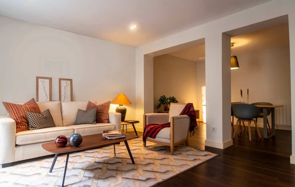Cozy living room featuring a beige sofa with decorative pillows, a wooden coffee table, and a stylish armchair, illuminated by a warm lamp, with a dining area visible in the background.