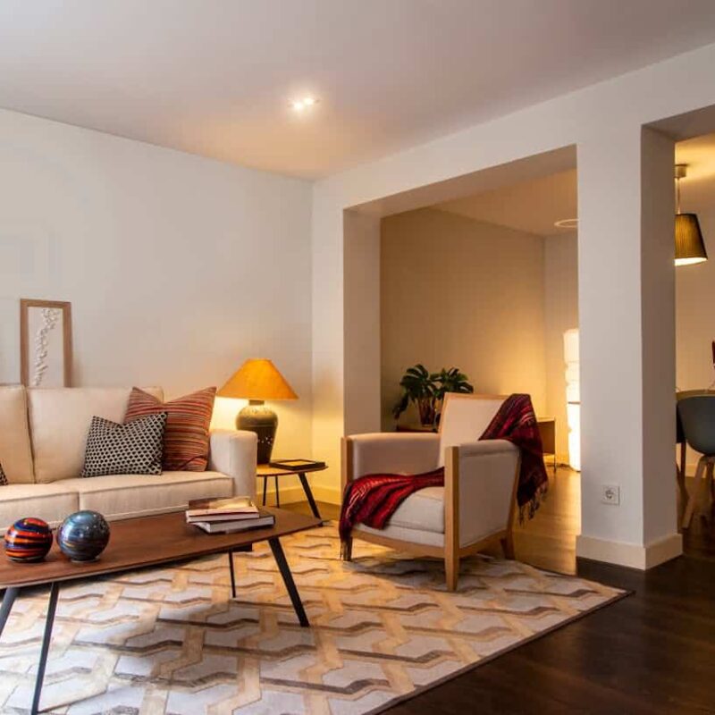 Cozy living room setup featuring a beige sofa with patterned cushions, a wooden coffee table displaying decorative objects, an accent lamp, and a potted plant, illustrating a welcoming home environment relevant to home buying and real estate discussions.